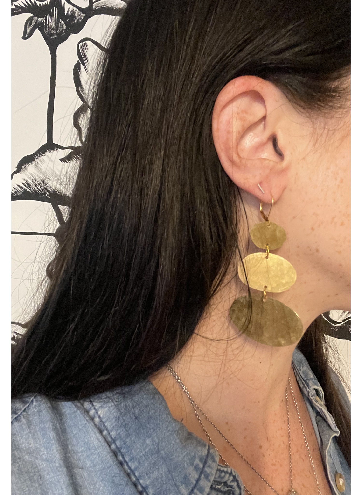 A close-up side profile of a person wearing a large, three-tiered gold dangle earring with organic, wavy disc shapes. The person has long dark hair and is wearing a light blue denim shirt.